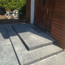 Schody wejściowe wyłożone szarą kostką brukową w kształcie prostokątów, prowadzące do drewnianych drzwi; w tle dekoracyjny rower z koszykami kwiatów.