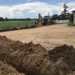 Prace ziemne w Giżycku: koparka przygotowuje teren pod fundament, widoczny wykop i pracownik w pomarańczowej kamizelce.