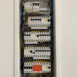 Rozdzielnica elektryczna z licznymi bezpiecznikami, przewodami w r&oacute;żnych kolorach i urządzeniem Urmet 1726/100, zamontowana w białej obudowie na ścianie.