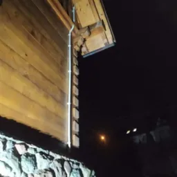 Drewniana ściana domu z kamienną podmurówką i białą rurą spustową, sfotografowana nocą z dołu.