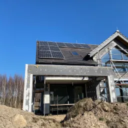 Dom w trakcie ocieplania elewacji z zamontowanymi panelami fotowoltaicznymi na dachu, widoczne rusztowania i przygotowany teren.