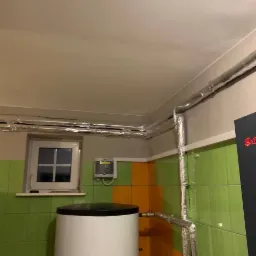 Biały bojler z czarną pokrywą w pomieszczeniu z zielono-pomarańczowymi kafelkami, widoczne rury w izolacji termicznej pod sufitem i przy ścianach.