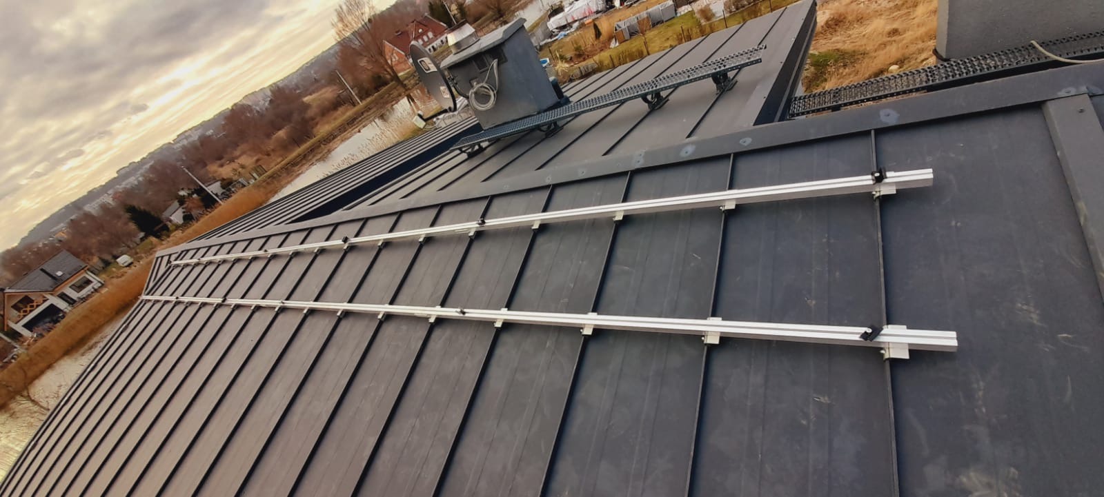 Montaż szyn pod panele słoneczne na nowym, ciemnym dachu z blachy trapezowej, widok z góry na tle zabudowań i pochmurnego nieba.
