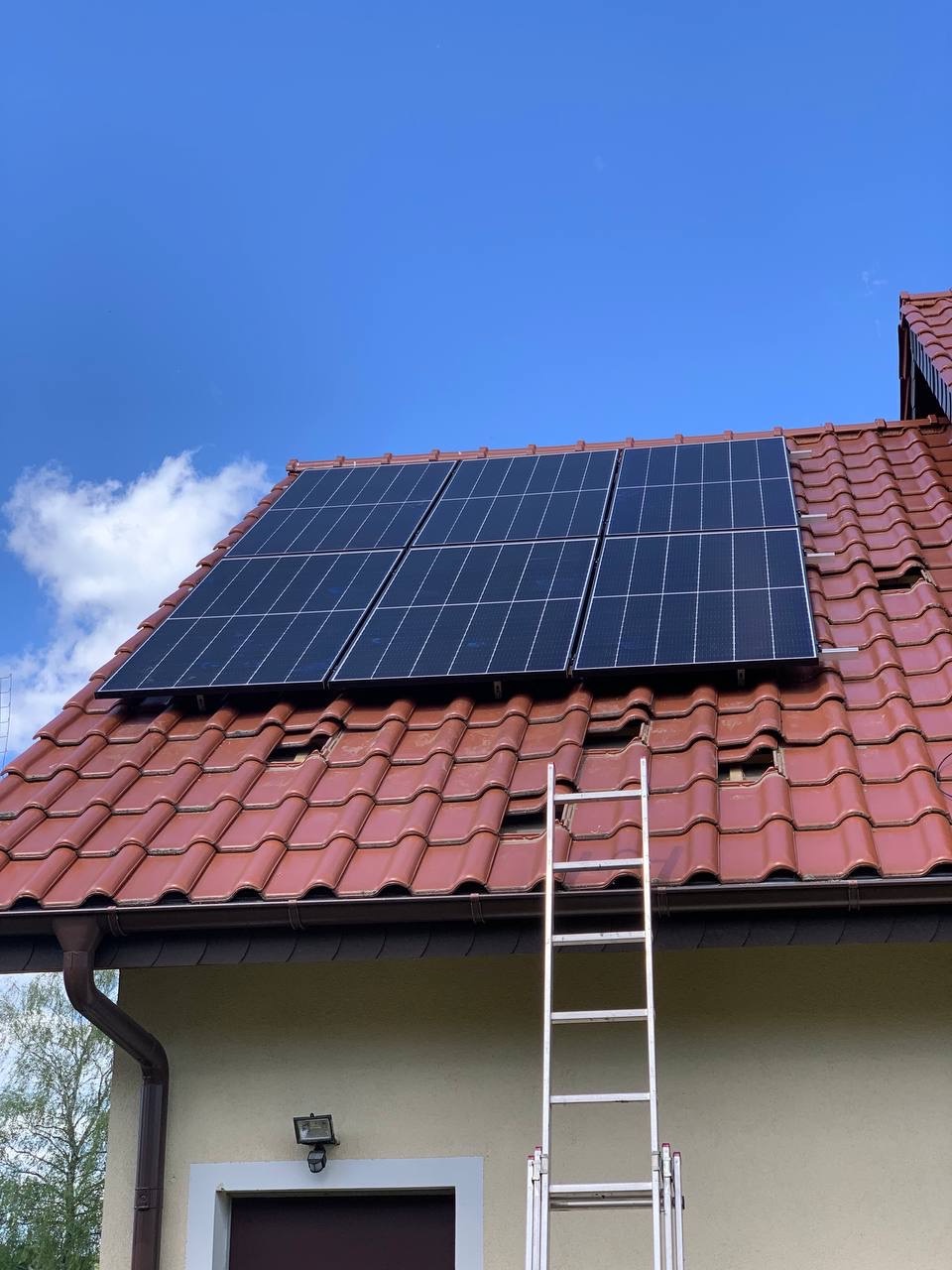 Panele słoneczne zamontowane na dachu z czerwonej dachówki, obok drabina oparta o rynnę i ścianę budynku, błękitne niebo w tle.