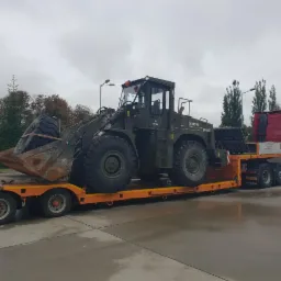 Transport niskopodwoziowy: Ciemnozielona ładowarka HSW SL-14C przewożona na pomarańczowej platformie przez czerwoną ciężarówkę Volvo, widok z boku na betonowym placu w pochmurny dzień.