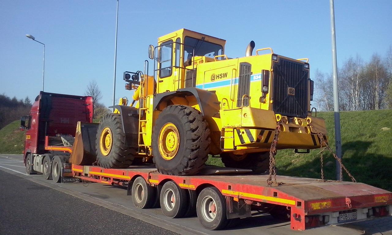 Żółta ładowarka kołowa HSW 534, zabezpieczona łańcuchami, przewożona na czerwonej niskopodwoziowej naczepie ciągniętej przez czerwonego Volvo FH na tle błękitnego nieba i zielonego wzgórza.