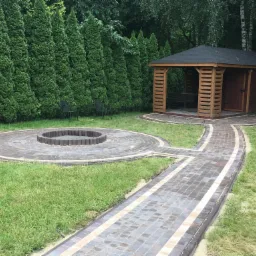 Ogród z altaną, ścieżką z kostki brukowej w odcieniach brązu i beżu, oraz okrągłym paleniskiem otoczonym kostką, na tle wysokich, zielonych tuj.