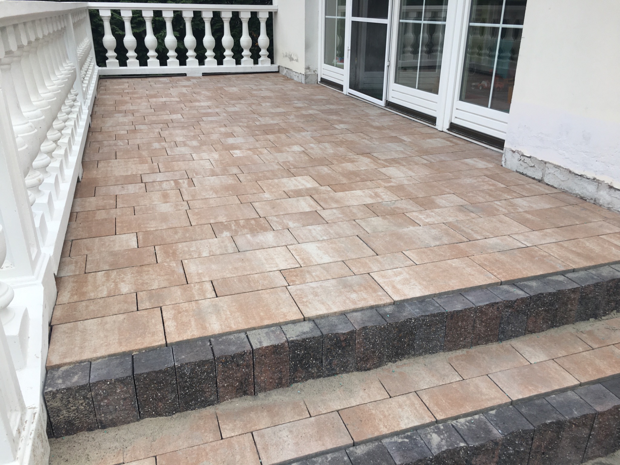 Wyłożony kostką taras z dwoma stopniami obramowanymi ciemniejszą kostką, biała balustrada w tle, widoczne drzwi balkonowe.