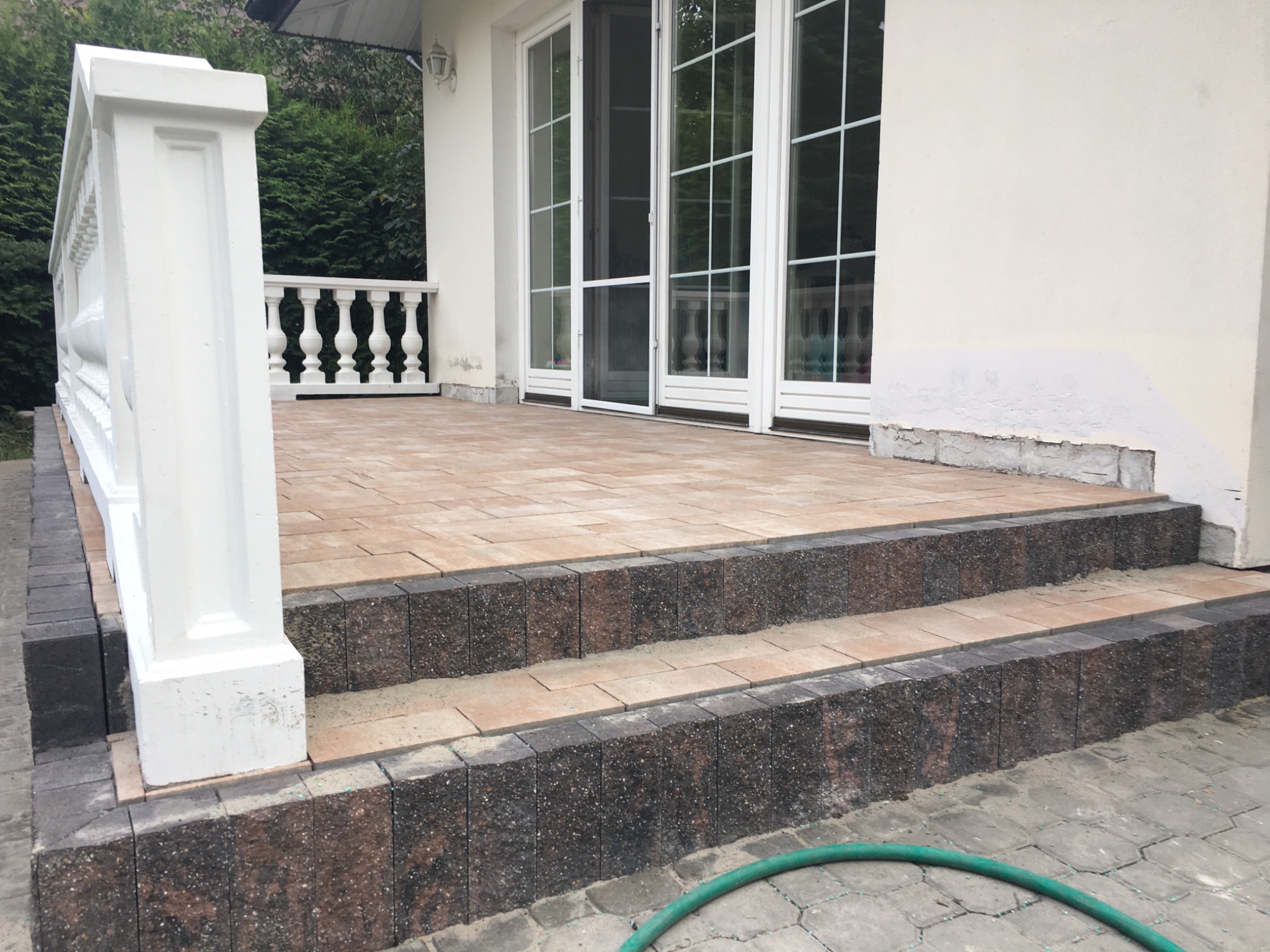 Wykończony taras z kostki brukowej w odcieniach beżu i brązu, z dwoma stopniami obłożonymi ciemniejszą kostką, biała balustrada po lewej stronie, widoczne drzwi tarasowe i fragment węża ogrodowego...