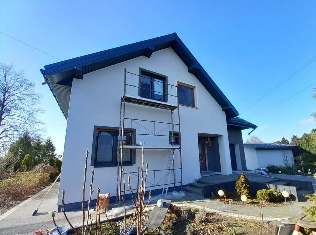 Dom jednorodzinny z szarą elewacją i ciemnymi ramami okiennymi w trakcie prac wykończeniowych, widoczne rusztowanie i niezabezpieczone okno bez ramy.