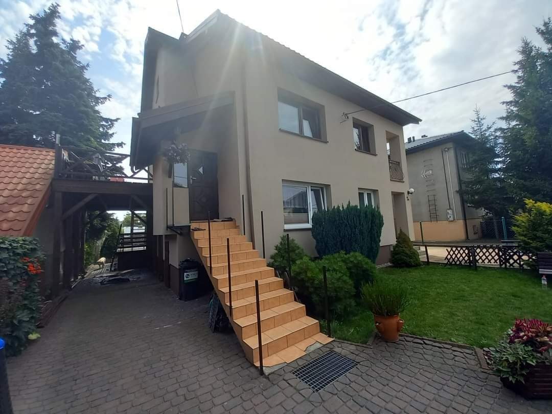 Wykończona elewacja domu jednorodzinnego w Gałkówku z zewnętrznymi schodami wejściowymi, widok z poziomu chodnika, jasna kolorystyka budynku.
