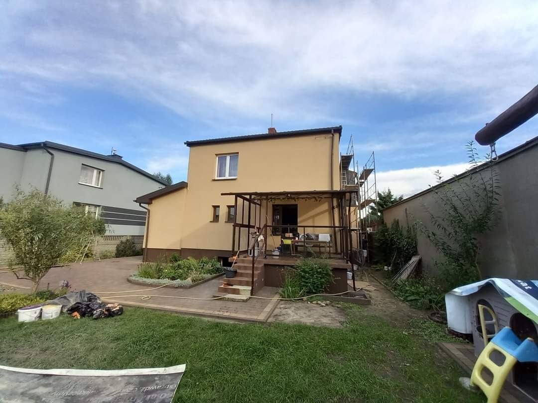 Dwukondygnacyjny dom w trakcie prac elewacyjnych, widoczne rusztowania, świeżo pomalowana fasada w odcieniach beżu, uporządkowany ogród z elementami placu zabaw.