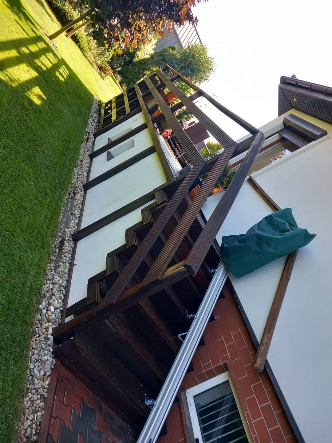 Drewniane schody zewnętrzne z balustradą, prowadzące do elewacji budynku z oknem, widoczne kamienie ozdobne i trawnik.