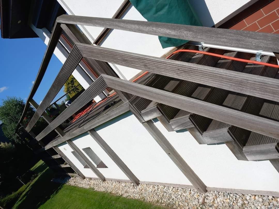 Boczne ujęcie zewnętrznych schodów z drewnianą balustradą, prowadzących do wejścia budynku z białą elewacją i zielonym zadaszeniem nad drzwiami.