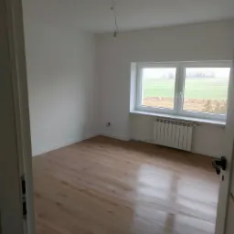 Pusty pokój z panelami podłogowymi o wzorze drewna, świeżo pomalowanymi białymi ścianami i grzejnikiem pod oknem z widokiem na zielone pole w pochmurny dzień.