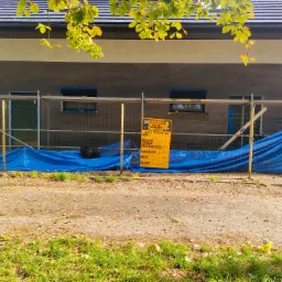 Budynek w trakcie prac elewacyjnych, zabezpieczony rusztowaniem i niebieską plandeką, widoczna tablica informacyjna budowy, jesienne liście na pierwszym planie.