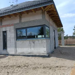 Niedokończony dom z białego bloczka z zamontowanymi oknami PCV z czarnymi ramami, widoczne drewniane elementy pod dachem i piasek wokół budynku.