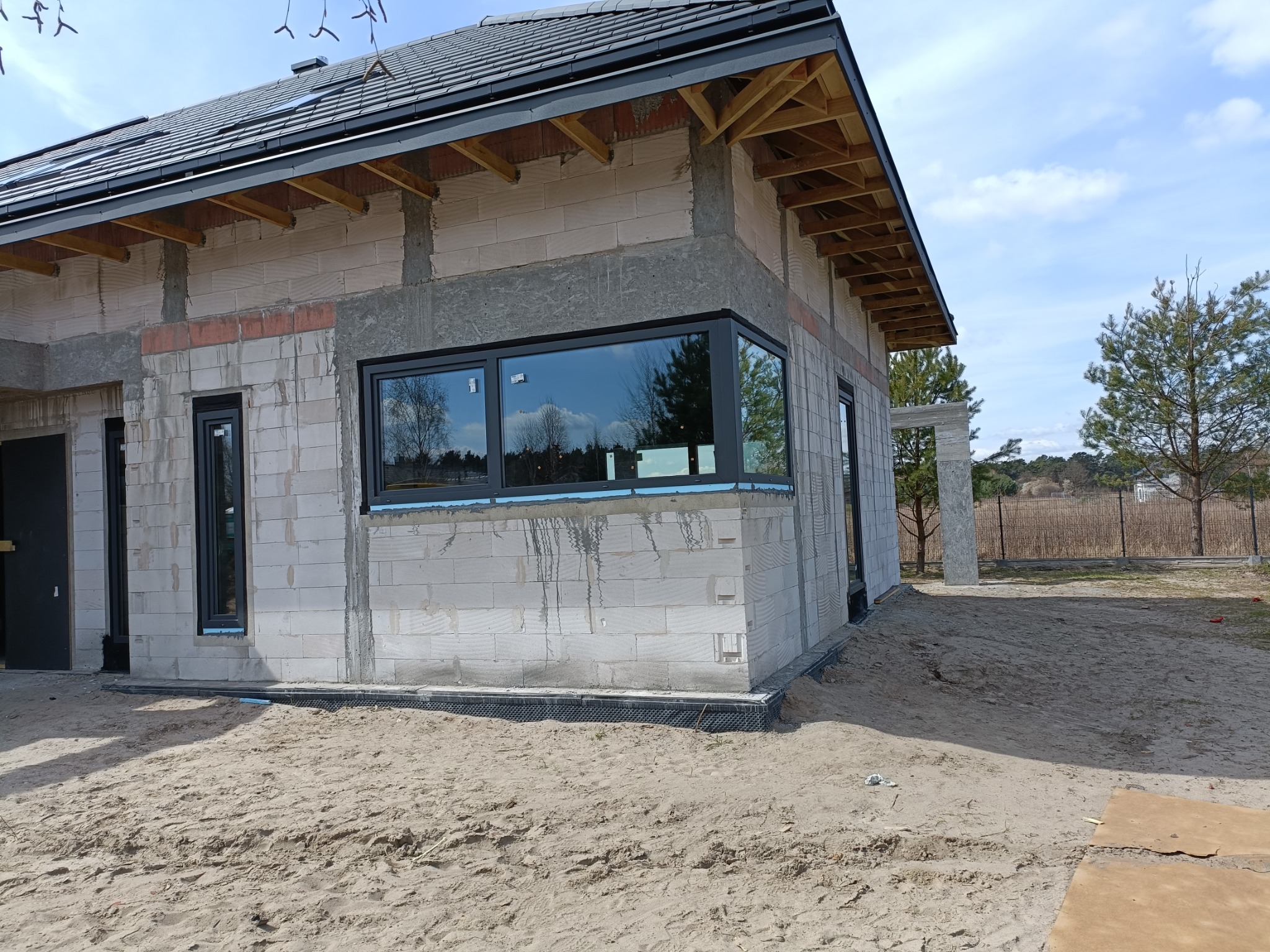 Niedokończony dom z białego bloczka z zamontowanymi oknami PCV z czarnymi ramami, widoczne drewniane elementy pod dachem i piasek wokół budynku.