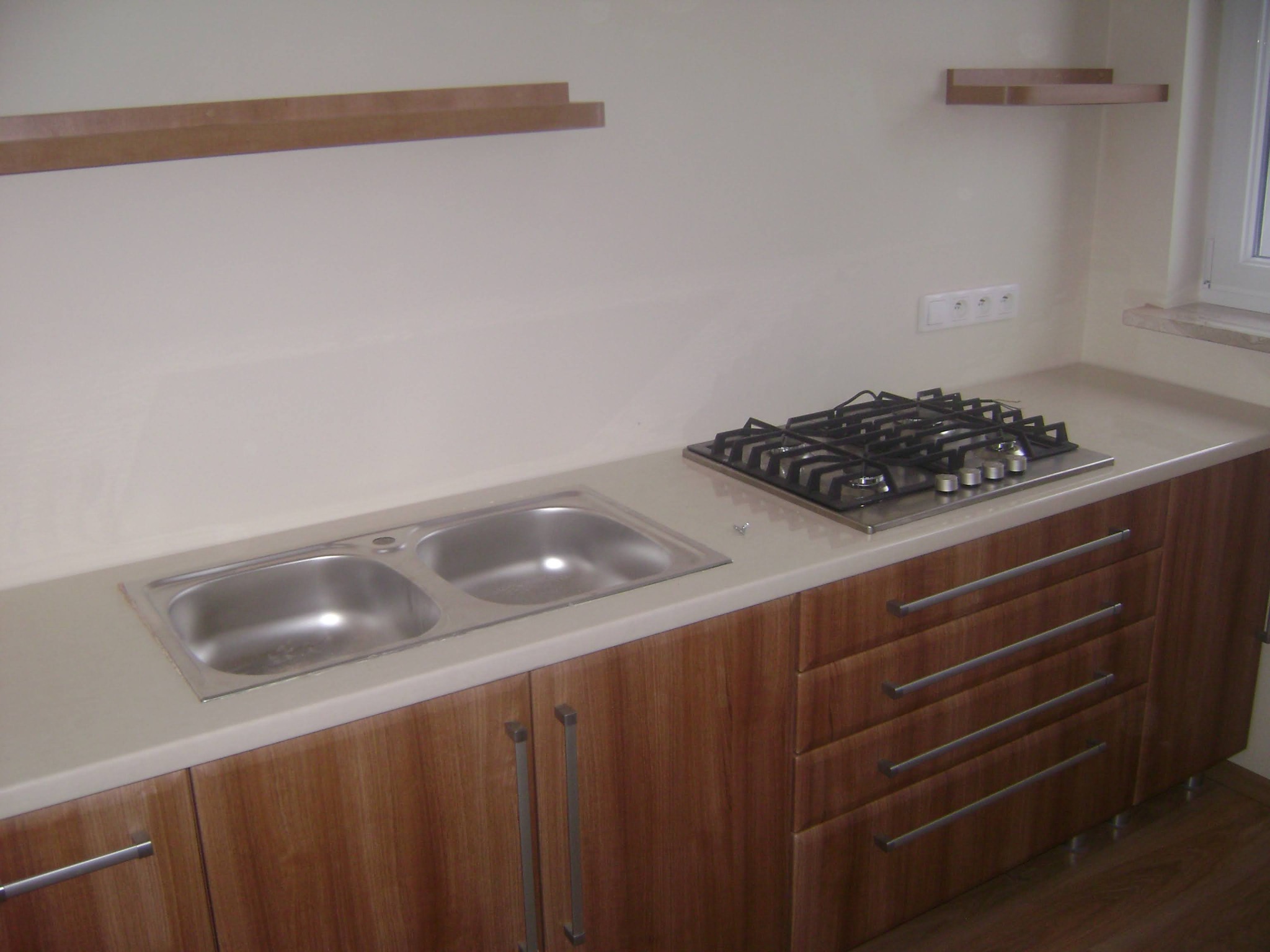 Fragment zabudowy kuchennej z dwukomorowym zlewozmywakiem i płytą gazową, fronty szafek w odcieniach brązu z poziomymi, metalowymi uchwytami, nad blatem dwie półki.