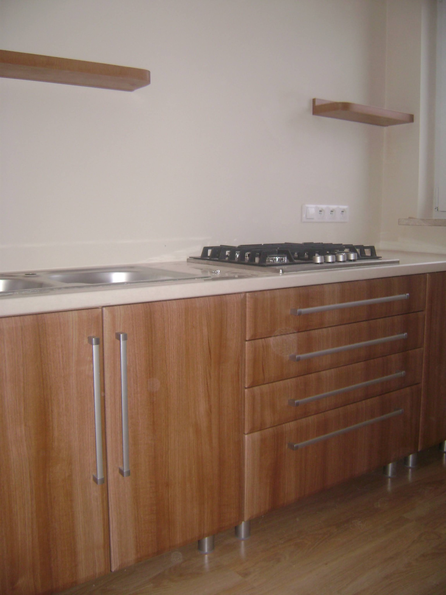 Fragment zabudowy kuchennej z drewnianymi frontami, widoczny zlew dwukomorowy i płyta gazowa na blacie, dwie półki na ścianie.