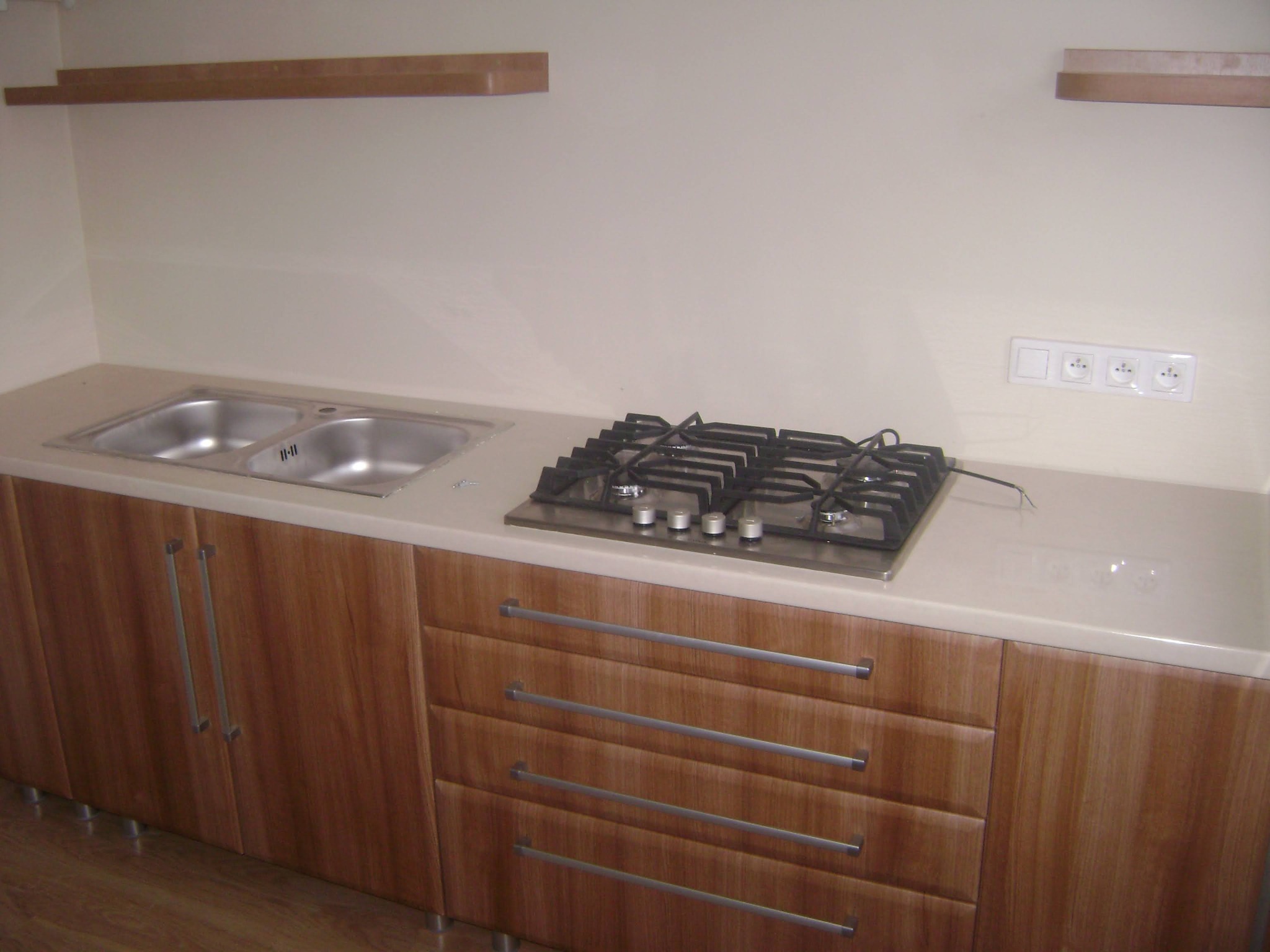 Fragment zabudowy kuchennej z drewnopodobnymi frontami, zlewem dwukomorowym i płytą gazową, widoczne gniazdka elektryczne na ścianie.