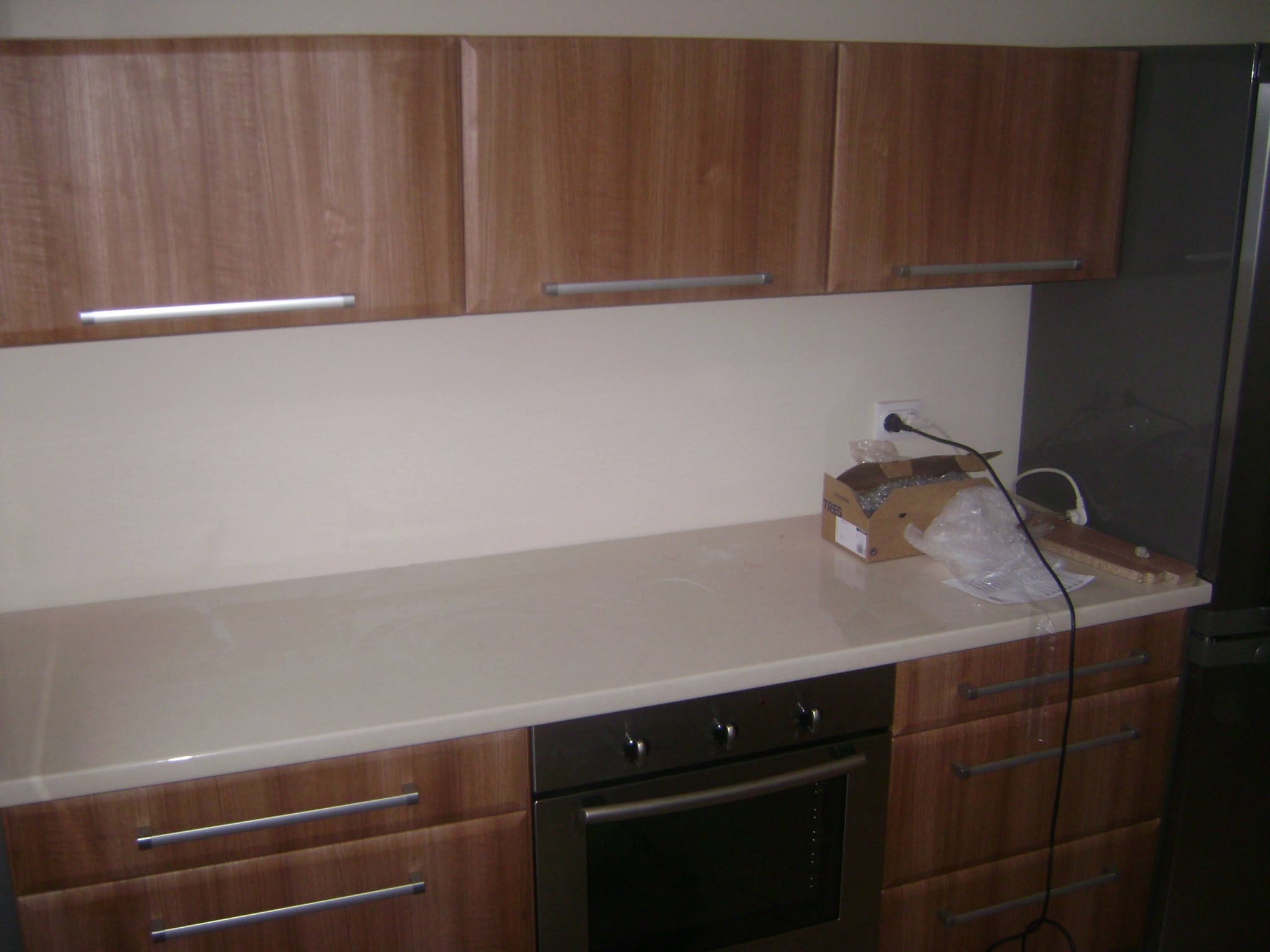 Fragment kuchni z drewnianymi szafkami górnymi i dolnymi z podłużnymi, srebrnymi uchwytami, jasnym blatem, wbudowanym piekarnikiem i elementami instalacji elektrycznej na blacie.