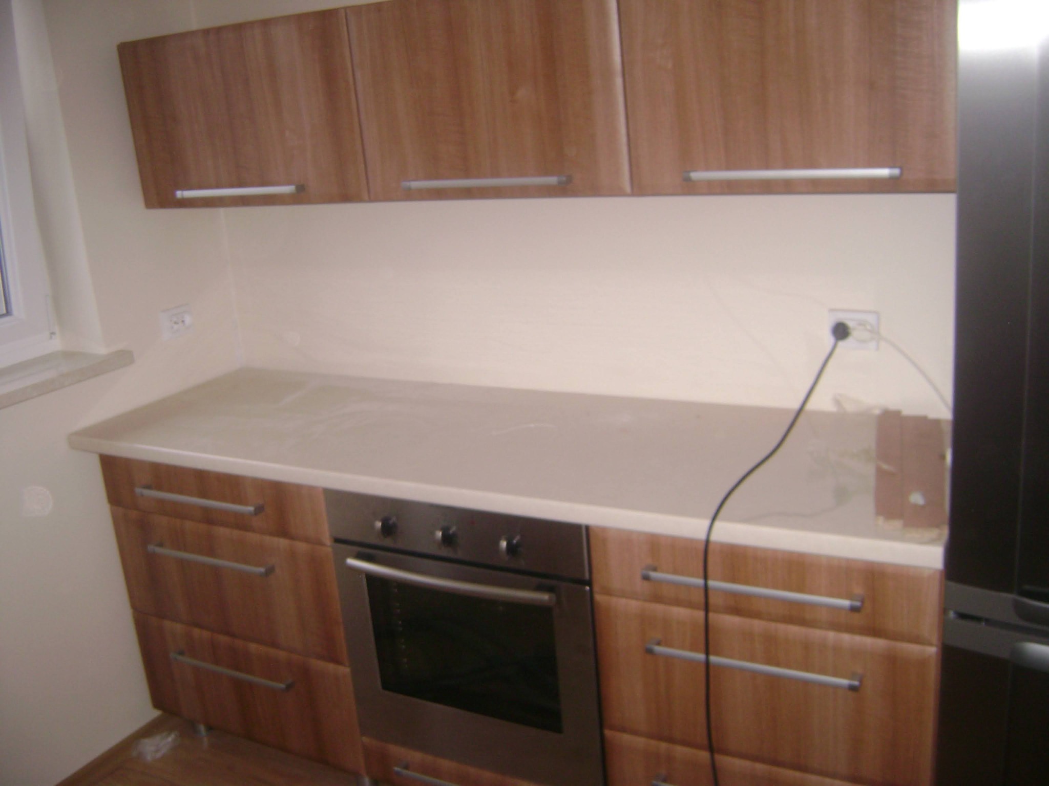 Fragment zabudowy kuchennej z drewnopodobnymi frontami, srebrnymi uchwytami, piekarnikiem i blatem w jasnym odcieniu, widoczne gniazdko elektryczne i kabel zasilający.