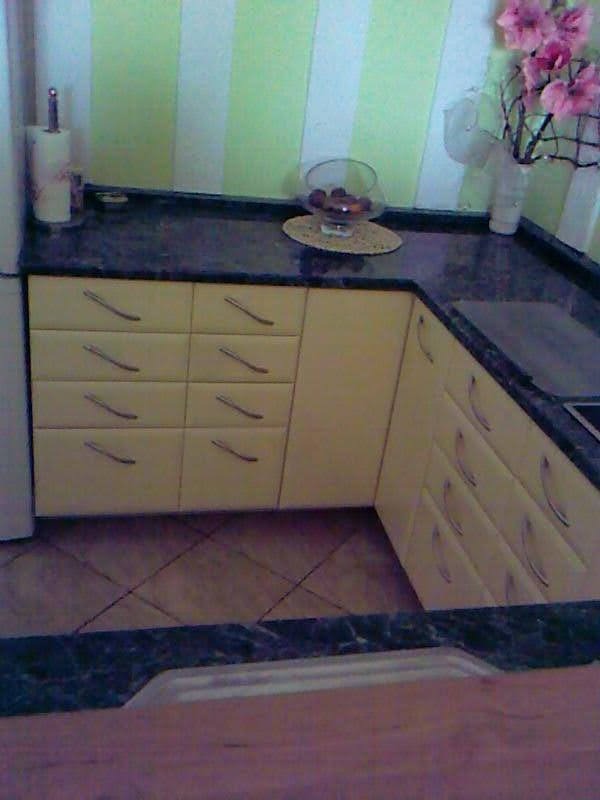 Żółta kuchnia na wymiar z ciemnym blatem i srebrnymi uchwytami szuflad, widok z niskiej perspektywy, na blacie szklana misa z owocami i kwiaty w wazonie.