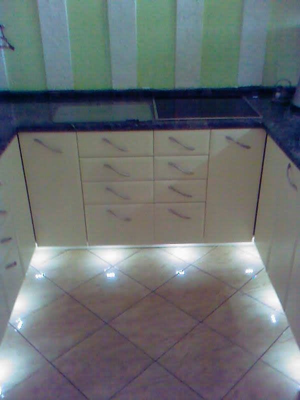 Fragment kremowej zabudowy kuchennej z ciemnym blatem i podświetleniem LED pod szafkami, widok na dolne szafki i część płyty grzewczej.