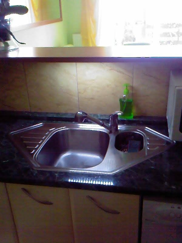 Narożny zlew ze stali nierdzewnej w blacie z ciemnego granitu, z widocznym dozownikiem na płyn do naczyń i ociekaczem, nad szafkami kuchennymi w odcieniach beżu.