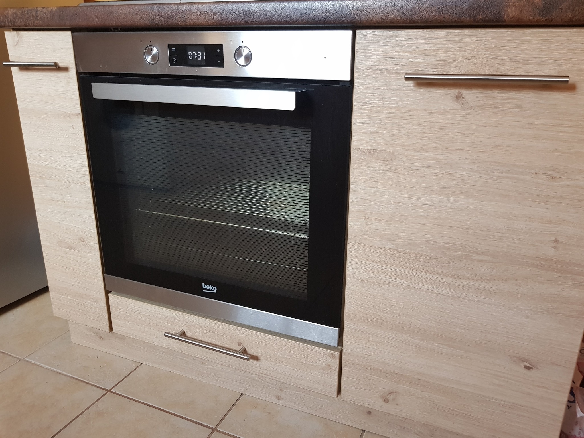 Wbudowany piekarnik marki Beko w szafce kuchennej z drewnopodobnym wykończeniem w Chodzieży, widoczne metalowe uchwyty i fragment blatu.