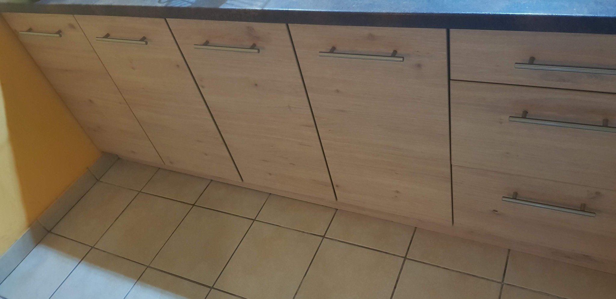 Fragment zabudowy kuchennej z jasnego drewna z poziomymi, metalowymi uchwytami, widoczny na tle beżowej podłogi z płytek.