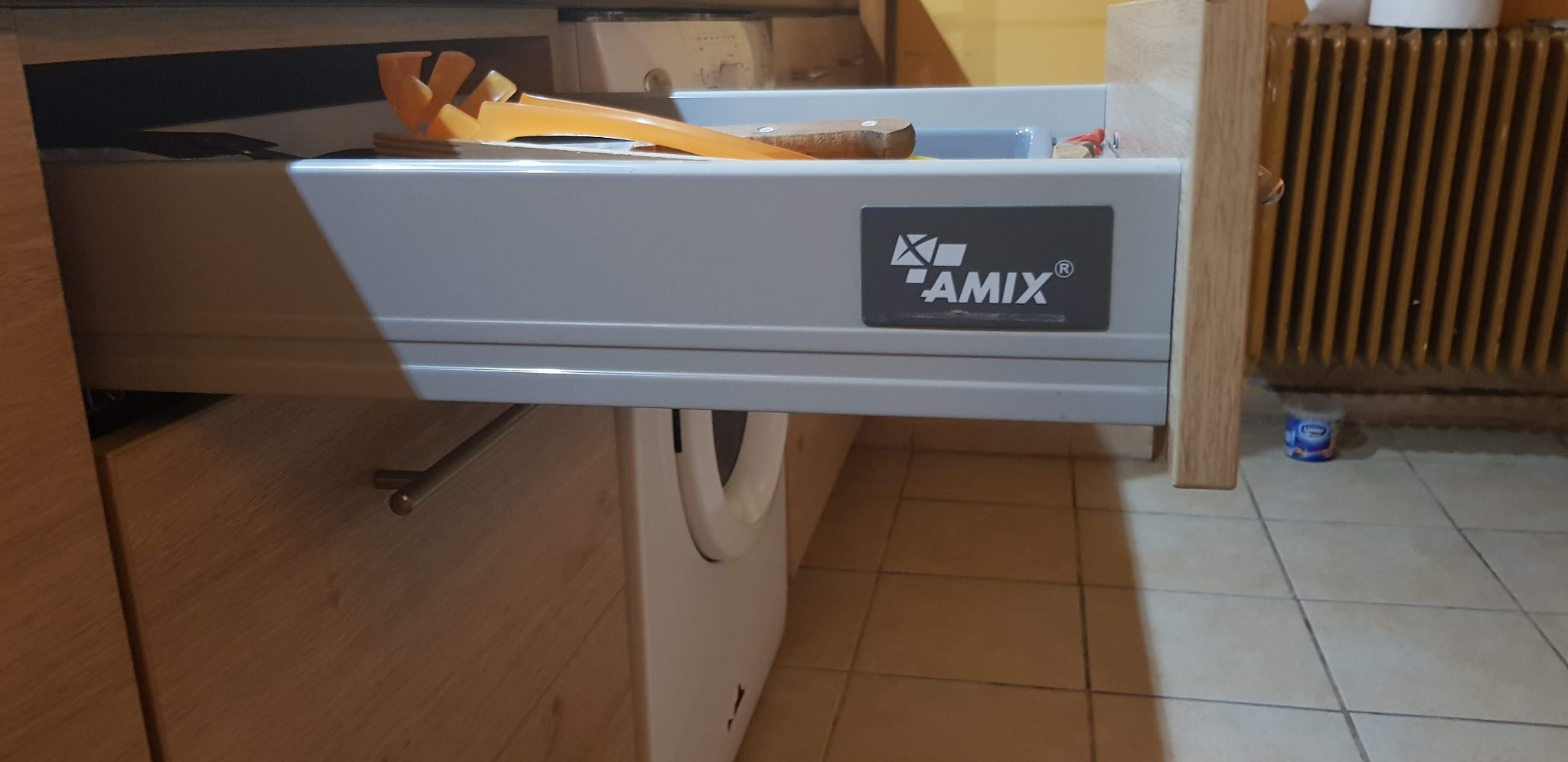 Otwarta szuflada kuchenna z logo AMIX, widoczne przybory kuchenne i fragment pralki.