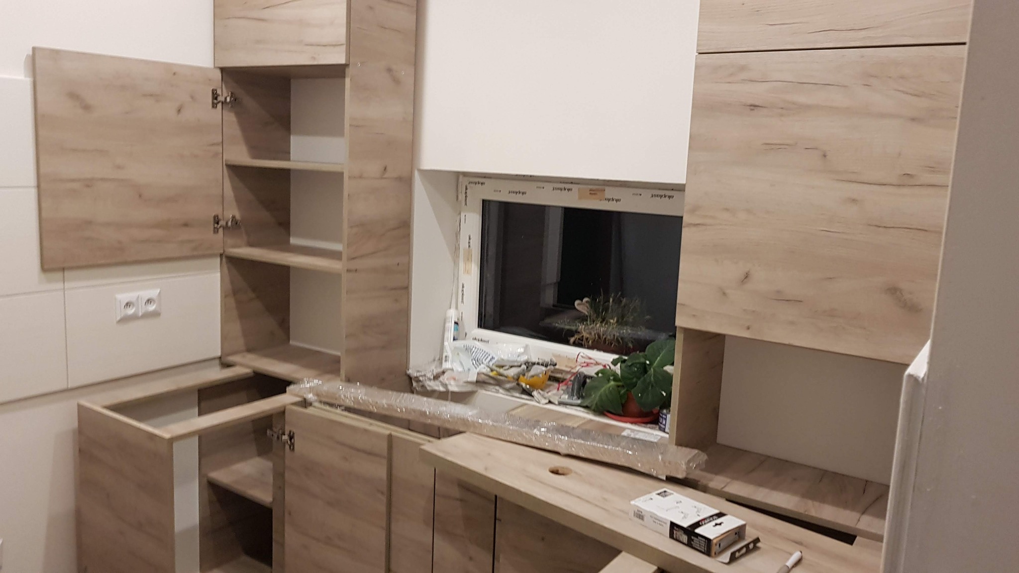 Montaż kuchni: surowe drewniane szafki kuchenne z otwartymi drzwiczkami i widocznymi półkami, okno z roślinami w tle, materiały budowlane i narzędzia na blacie.