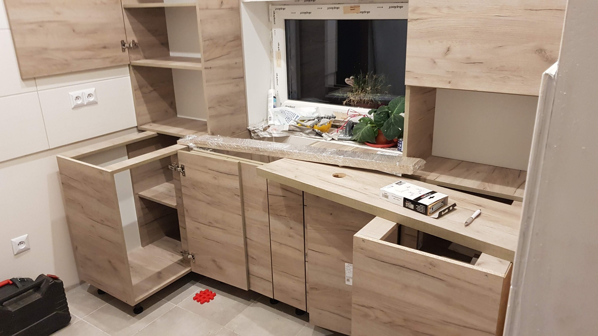 Montaż szafek kuchennych z drewnopodobnym wykończeniem, widoczne elementy w trakcie instalacji, narzędzia i akcesoria na blacie, okno w tle.