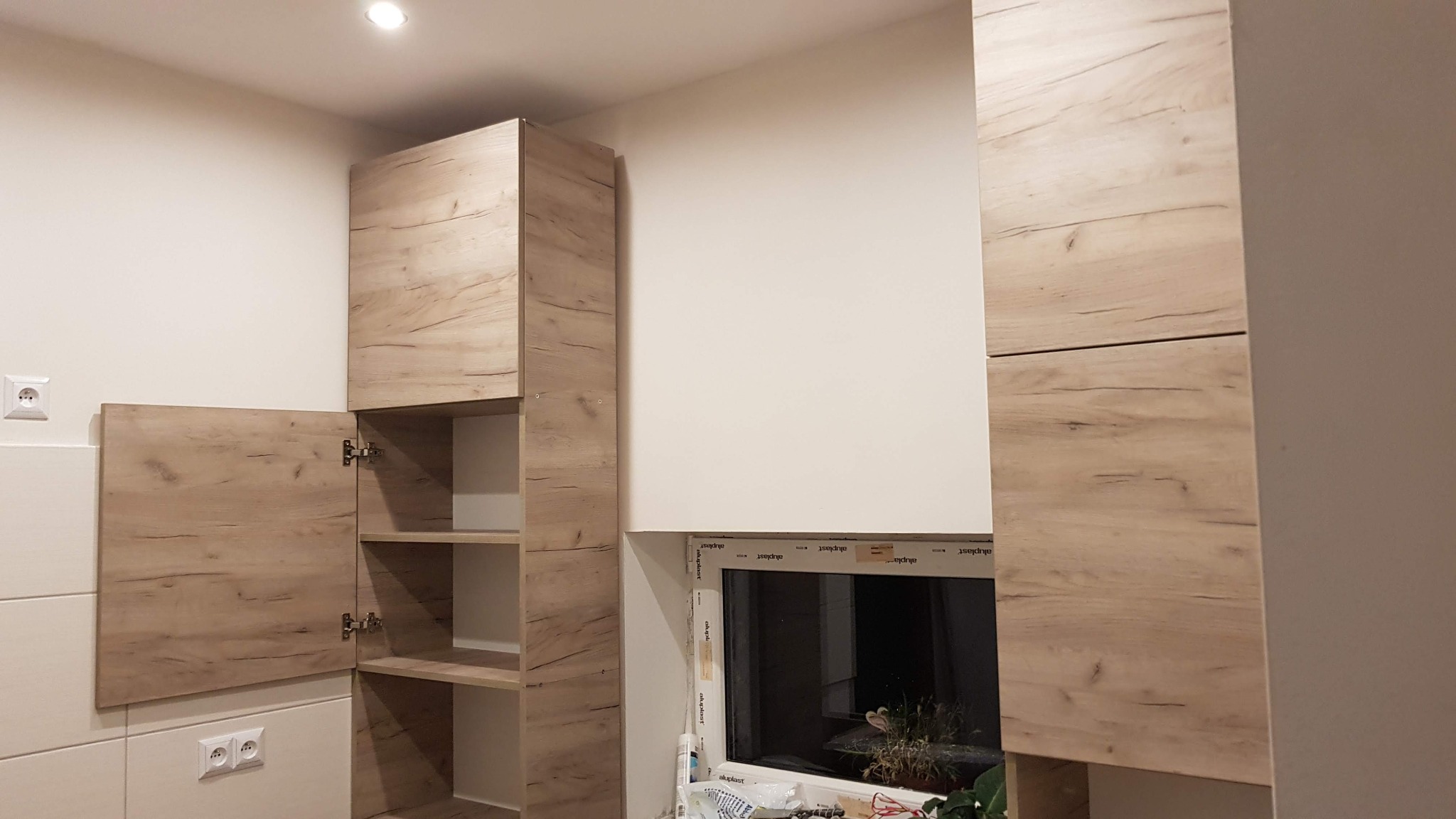 Nowoczesna zabudowa kuchenna z drewnopodobnymi frontami, widoczne gniazdka elektryczne, otwarte szafki z półkami, okno z rośliną w tle.