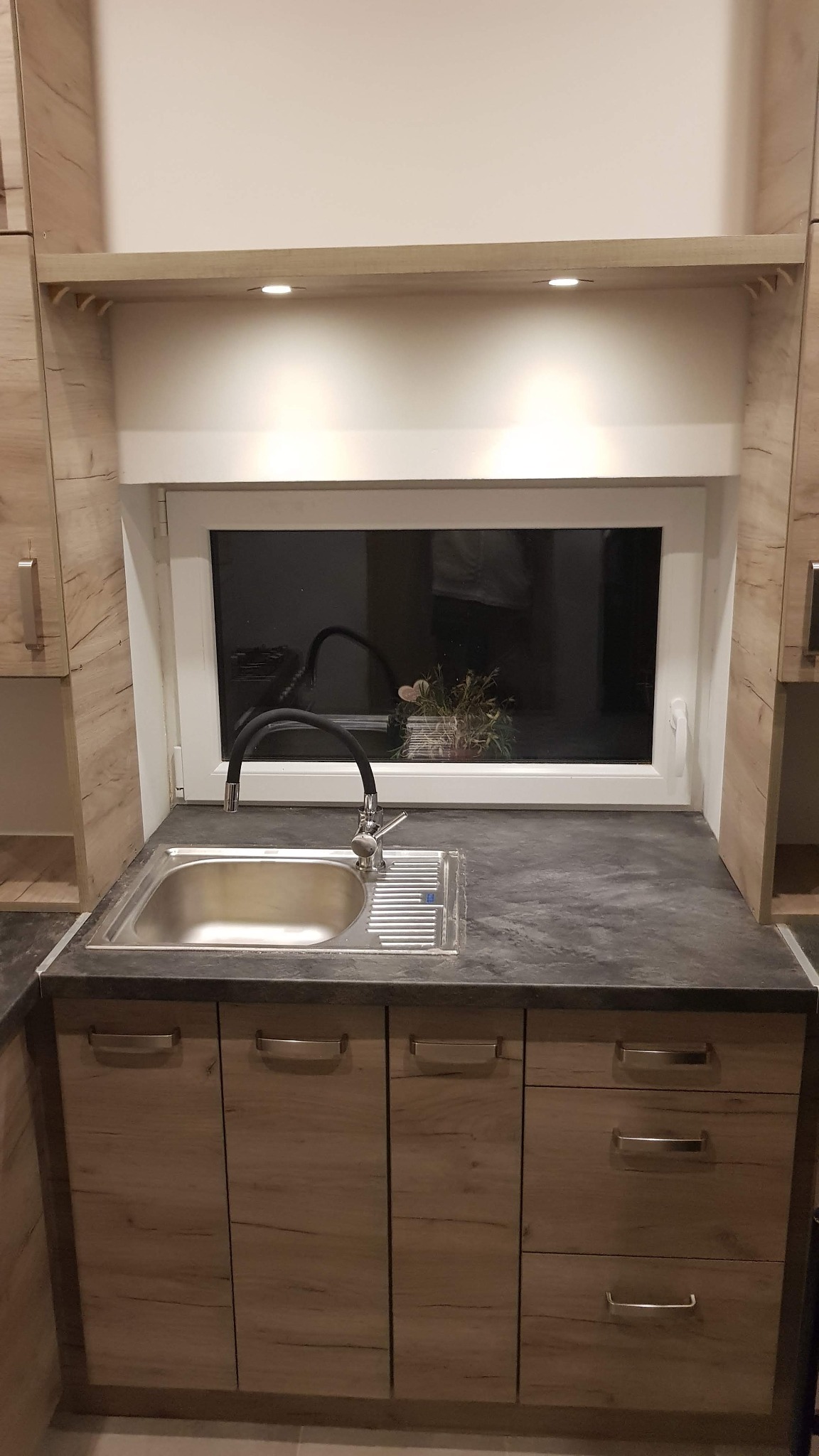 Nowoczesna kuchnia z drewnianymi szafkami, stalowym zlewem i czarną baterią, oświetlona punktowo pod półką nad oknem.