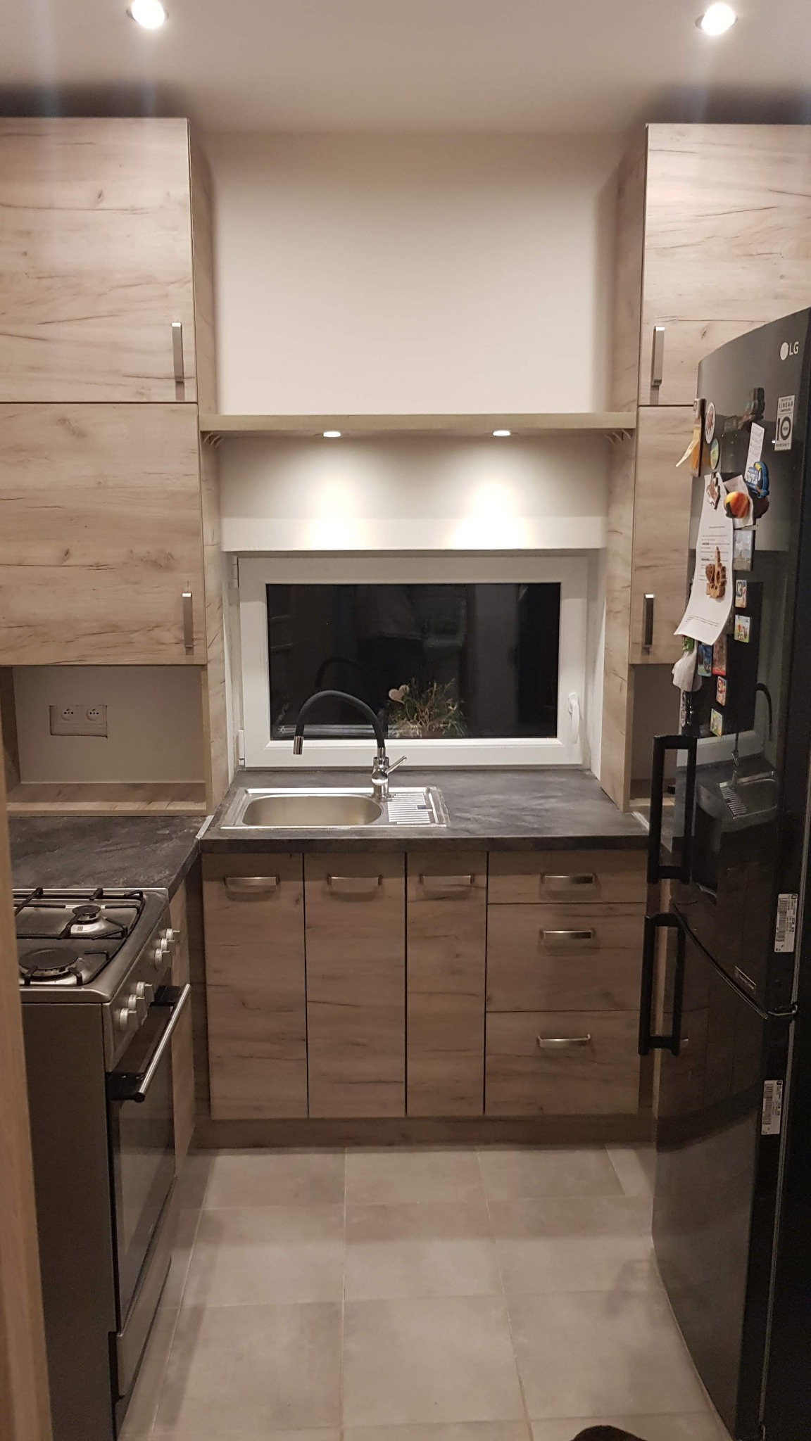 Nowoczesna kuchnia z drewnianymi frontami, czarnym blatem i oknem nad zlewem, oświetlenie punktowe pod szafkami wiszącymi, widoczna płyta gazowa i lodówka.