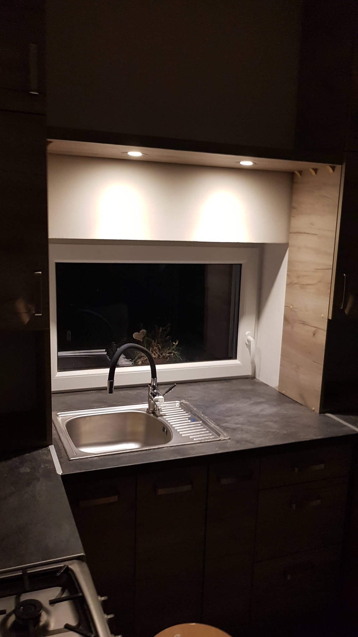 Nowoczesna kuchnia z ciemnymi szafkami i blatem, stalowy zlew z czarną baterią na tle okna z widokiem na zieleń, oświetlenie punktowe pod szafkami wiszącymi.