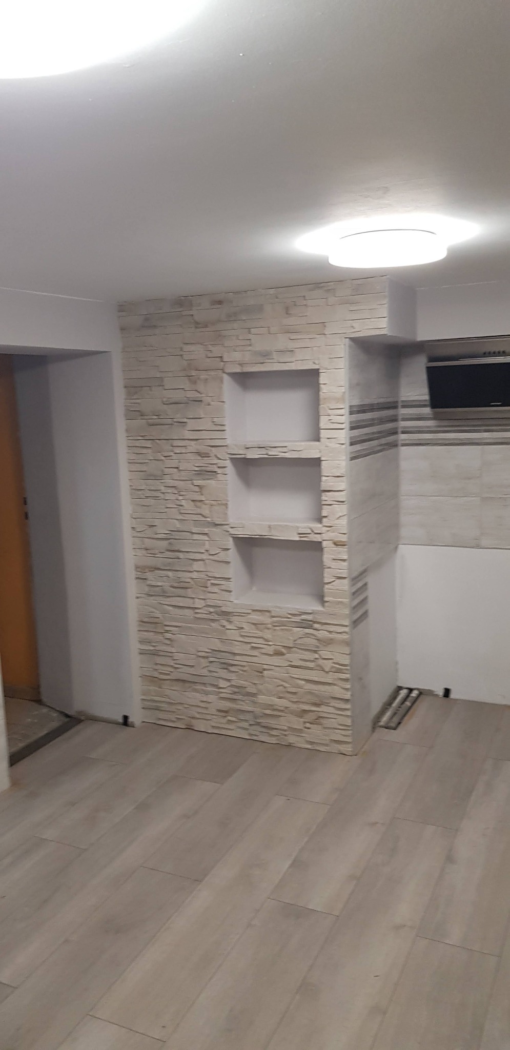 Ściana wykończona dekoracyjną okładziną kamienną z wnękami na półki, sąsiadująca z fragmentem zabudowy kuchennej i jasną podłogą z paneli.