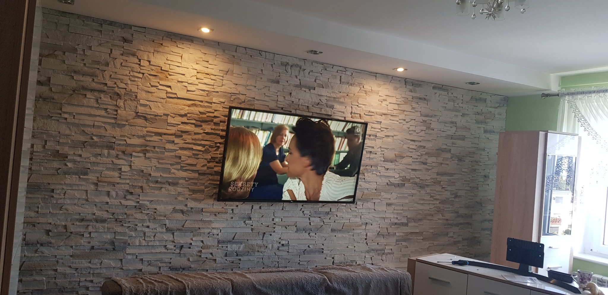 Ściana z dekoracyjnej szarej cegły z zamontowanym telewizorem. Na szafce pod telewizorem leżą narzędzia.