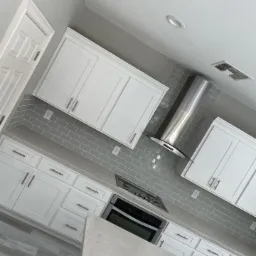 Nowoczesna kuchnia z białymi szafkami, szarymi płytkami metra i stalowym okapem, widziana pod kątem.