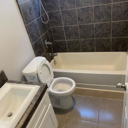 Scleaningkrk - Łazienka w Krakowie po remoncie: widok na białą wannę, umywalkę z szarą marmurową płytą i toaletę z otwartą klapą, szare płytki na ścianach i podłodze.