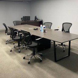 Scleaningkrk - Sala konferencyjna z długim, drewnianym stołem, krzesłami biurowymi na kółkach, biurkiem z komputerem w tle oraz butelkami płynów do dezynfekcji na stole.