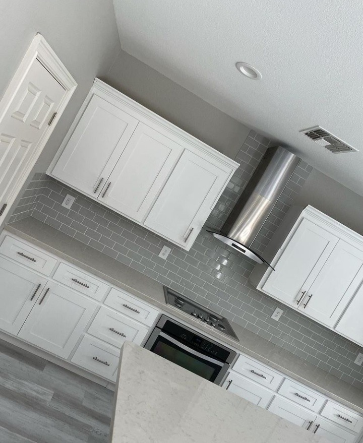 Nowoczesna kuchnia z białymi szafkami, szarymi płytkami metra i stalowym okapem, widziana pod kątem.