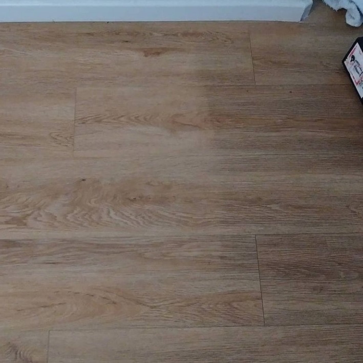 Panel podłogowy imitujący drewno, widoczne zabrudzenia wzdłuż krawędzi paneli i przy listwie przypodłogowej.