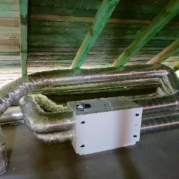 Biała jednostka rekuperacyjna z podłączonymi, izolowanymi przewodami wentylacyjnymi, zamontowana na poddaszu z drewnianą konstrukcją dachu.