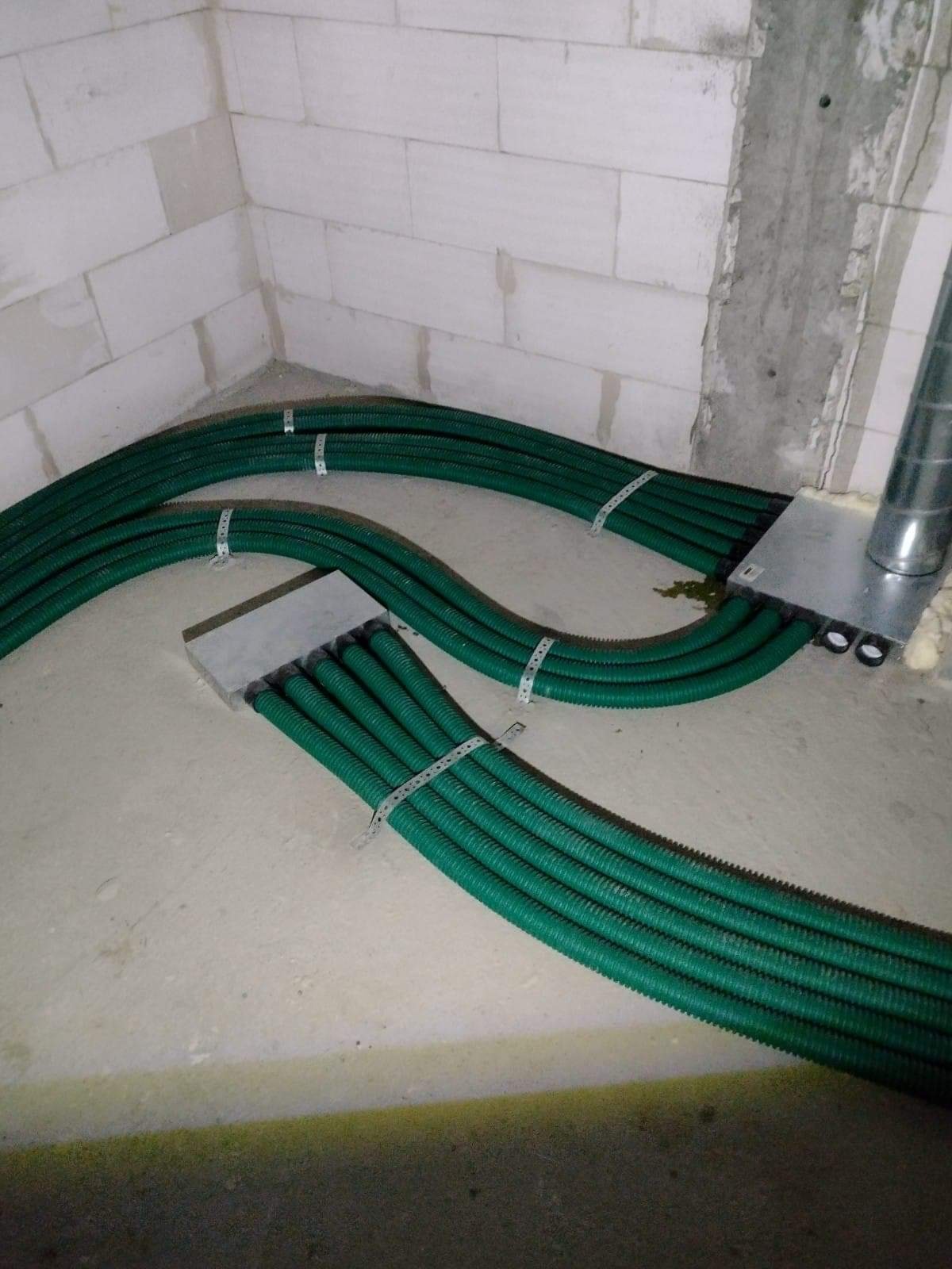 Zielone, karbowane rury wentylacyjne prowadzone wzdłuż ściany z betonu komórkowego, zabezpieczone metalowymi opaskami zaciskowymi, wchodzące do metalowej skrzynki rozdzielczej z izolacją.