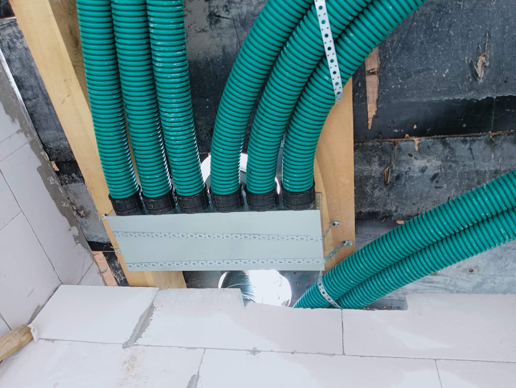 Zielone, karbowane rury wentylacyjne prowadzone przez drewniane belki stropowe, przymocowane perforowaną taśmą montażową, widoczne białe bloczki konstrukcyjne.