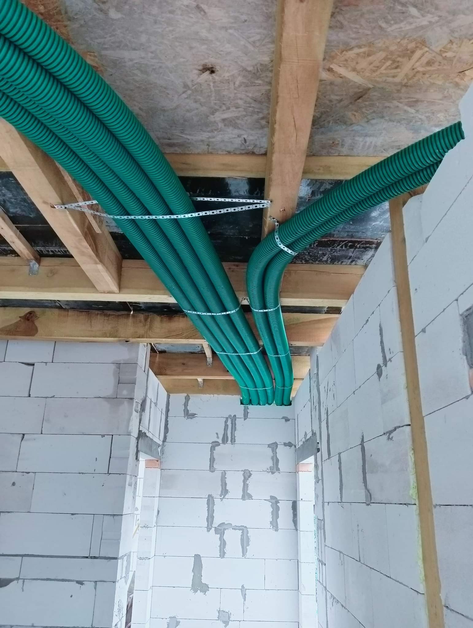 Zielone, karbowane rury wentylacyjne prowadzone pod drewnianym stropem w surowym wnętrzu z białych bloczków.